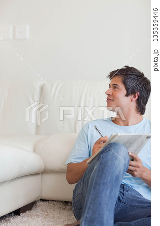 Man sitting in living room leaning against leather sofa holding pen and newspaper, copy space 135359446