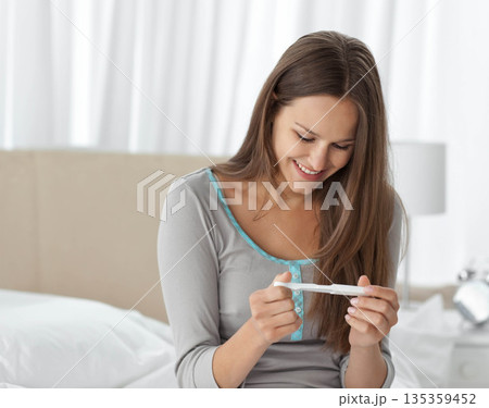 Woman examining pregnancy test while sitting on bed in softly lit bedroom with lamp, copy space 135359452