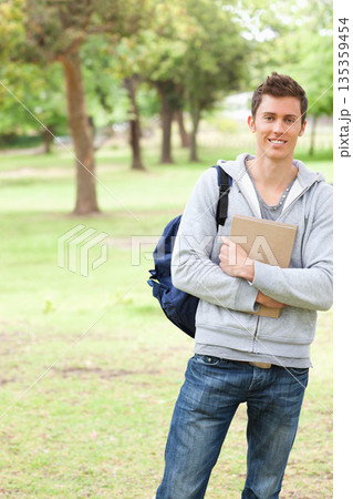 Male student standing in park holding hardcover notebook and carrying navy backpack, copy space Male student standing in park holding hardcover notebook and carrying navy backpack, copy space 135359454