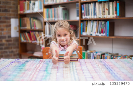 Female child student leaning on stacked books on patterned tablecloth in library giving thumbs up Female child student leaning on stacked books on patterned tablecloth in library giving thumbs up 135359470
