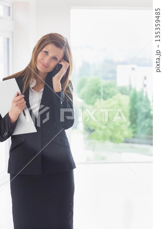Businesswoman holding smartphone and writing on clipboard with pen by office window, copy space Businesswoman holding smartphone and writing on clipboard with pen by office window, copy space 135359485