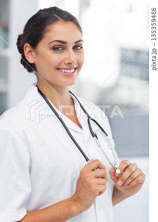 Woman wearing white lab coat holding stethoscope and smiling in medical clinic window light 135359486