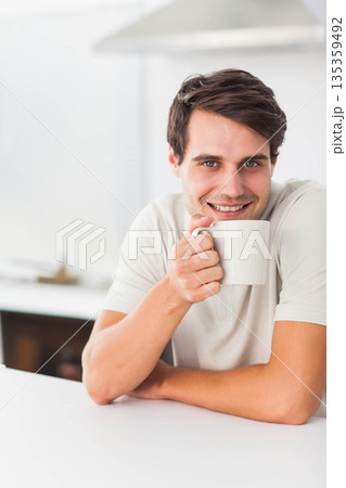 Man in his twenties sitting at kitchen counter holding white mug near mouth, smiling, copy space Man in his twenties sitting at kitchen counter holding white mug near mouth, smiling, copy space 135359492