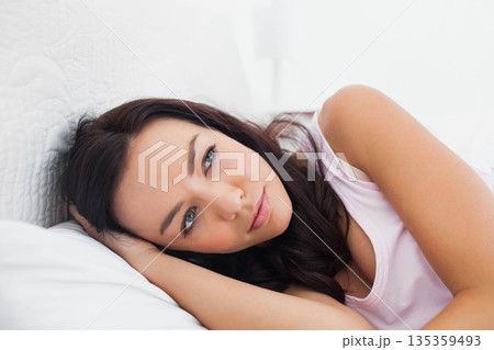 Woman lying on bed propped on side gazing thoughtfully in bedroom with white pillow and quilt Woman lying on bed propped on side gazing thoughtfully in bedroom with white pillow and quilt 135359493