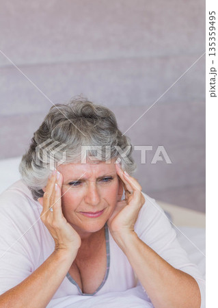 Senior woman leaning forward on bed pressing temples with pillow by pastel wall in pink sleepwear Senior woman leaning forward on bed pressing temples with pillow by pastel wall in pink sleepwear 135359495