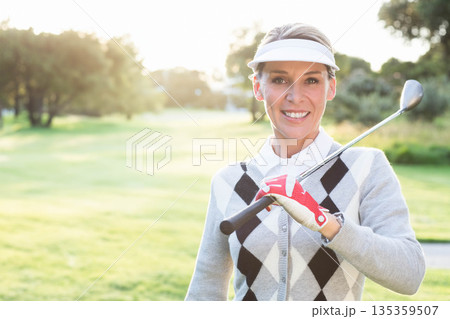 Middle-aged woman holding golf club across shoulders on golf fairway at sunset wearing visor, glove Middle-aged woman holding golf club across shoulders on golf fairway at sunset wearing visor, glove 135359507