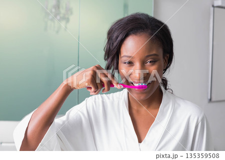 African American woman brushing teeth with pink toothbrush in bathroom wearing white bathrobe 135359508