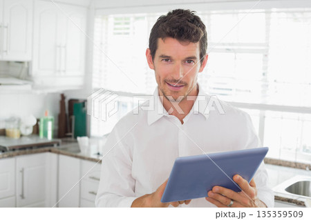 Man wearing button-up shirt using blue tablet standing in kitchen at granite countertop near sink Man wearing button-up shirt using blue tablet standing in kitchen at granite countertop near sink 135359509