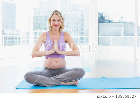 Pregnant woman practicing yoga in sportswear on blue mat under large windows at yoga studio 135359527