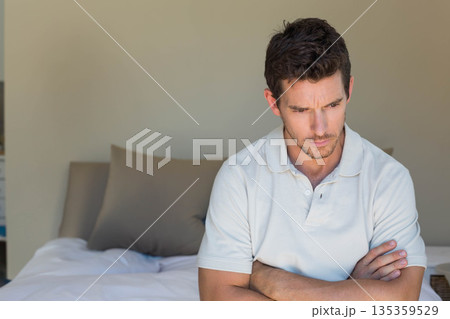 Man wearing white polo sitting on bed edge gazing in bedroom with taupe pillows, copy space 135359529