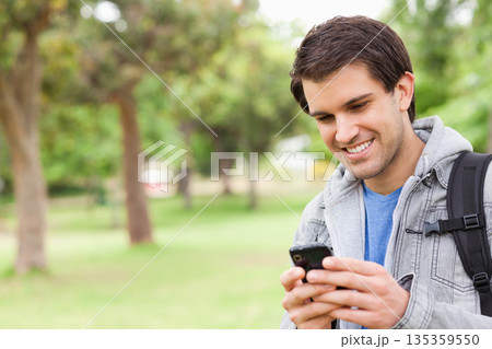 Man standing outdoors in lush green park tapping smartphone and carrying backpack, copy space 135359550