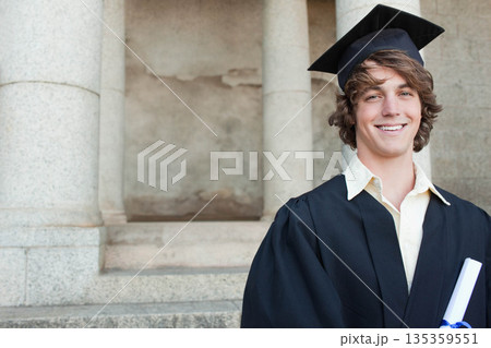 Male graduate holding diploma scroll standing by steps and columns in cap and gown, copy space 135359551
