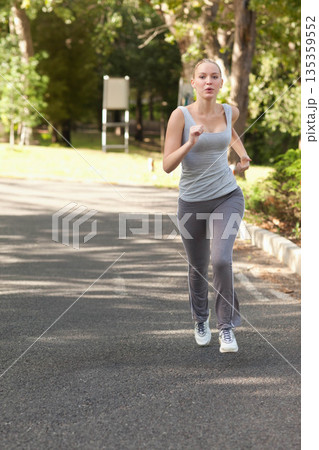 Female runner moving along sunlit tree-lined paved path in park wearing leggings and running shoes 135359552