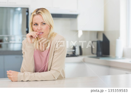 Middle-aged woman leaning forward touching lips and gazing left at kitchen countertop, copy space 135359583