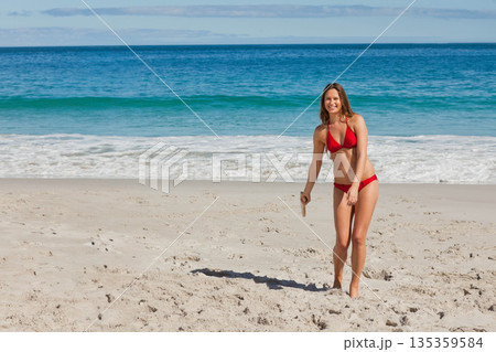Woman standing on sand near water edge wearing red bikini holding wooden boomerang, copy space 135359584