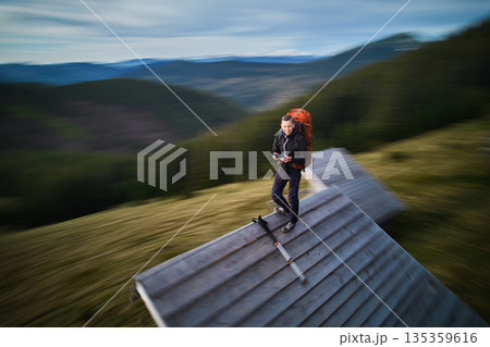 Hiker with orange backpack stands on roof of wooden cabin, surrounded by blurred, dynamic scenery. Motion blur effect. Hiker with orange backpack stands on roof of wooden cabin, surrounded by blurred, dynamic scenery. Motion blur effect. 135359616
