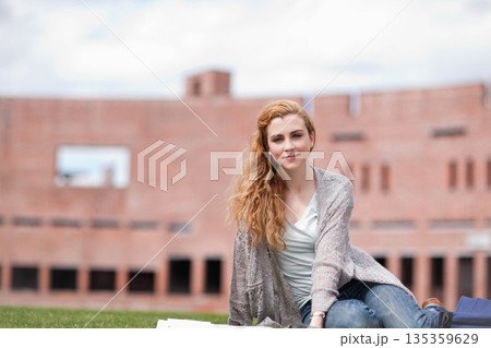 Female student sitting on lawn in front of brick building studying with open notebooks, copy space 135359629