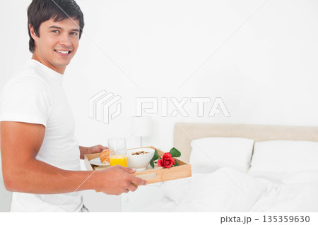 Man in twenties carrying tray with orange juice croissant rose to bed in bedroom, copy space 135359630