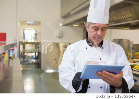 Senior chef in uniform holding tablet, entering data in kitchen with steel counters, copy space Senior chef in uniform holding tablet, entering data in kitchen with steel counters, copy space 135359647