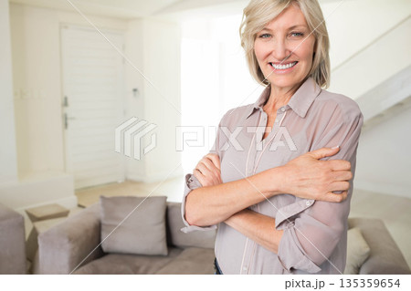Mature woman standing arms crossed smiling at camera in living room with sofa cushions, copy space Mature woman standing arms crossed smiling at camera in living room with sofa cushions, copy space 135359654