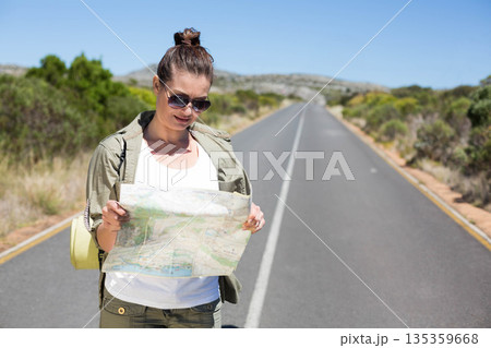Middle-aged woman standing on rural road holding paper map wearing backpack with mat, sunglasses 135359668