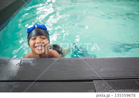African American boy emerging from pool, leaning on wood deck in black cap and blue goggles African American boy emerging from pool, leaning on wood deck in black cap and blue goggles 135359678