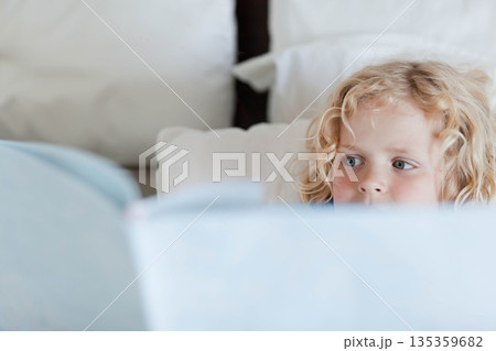Child boy reading pale blue hardcover book while lying on bed among white pillows, copy space Child boy reading pale blue hardcover book while lying on bed among white pillows, copy space 135359682