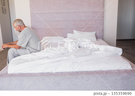 Senior man sitting on edge of unmade bed in pastel-toned bedroom with padded headboard, copy space Senior man sitting on edge of unmade bed in pastel-toned bedroom with padded headboard, copy space 135359690