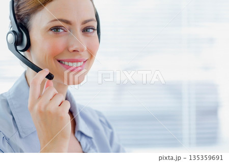 Smiling woman in collared shirt adjusting headset microphone at office with blinds, copy space 135359691
