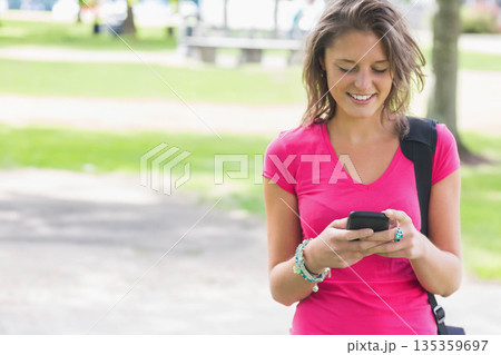 Woman in twenties wearing pink, jewelry, texting on smartphone with black bag in park, copy space Woman in twenties wearing pink, jewelry, texting on smartphone with black bag in park, copy space 135359697