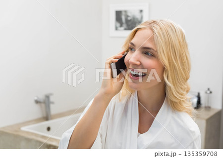 Woman standing at bathroom vanity wearing bathrobe talking on smartphone near sink, copy space 135359708