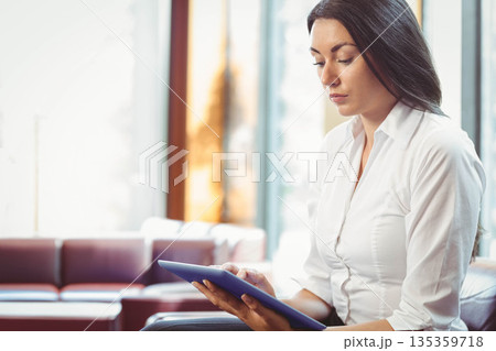Businesswoman reviewing tablet content while sitting on cushioned sofa in office lounge, copy space Businesswoman reviewing tablet content while sitting on cushioned sofa in office lounge, copy space 135359718