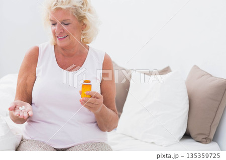 Senior woman sitting in bright bedroom holding amber pill bottle with white pills, copy space 135359725