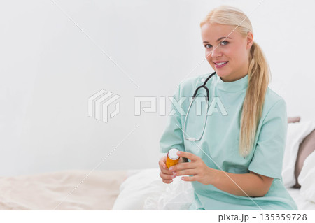 Adult woman professional in scrubs sitting on bed holding pill bottle with stethoscope, copy space Adult woman professional in scrubs sitting on bed holding pill bottle with stethoscope, copy space 135359728