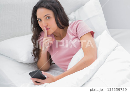 Woman wearing pink t-shirt reclining under white comforter holding smartphone shushing in bedroom 135359729