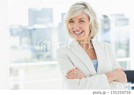 Middle-aged woman standing with arms crossed in office at window overlooking skyline, copy space 135359730
