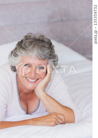 Senior woman smiling while reclining on bed with white sheets in bedroom showing ring on hand Senior woman smiling while reclining on bed with white sheets in bedroom showing ring on hand 135359732