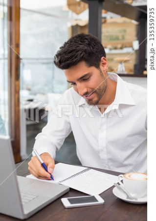 Man sitting at cafe table writing notes in notebook beside laptop smartphone and cappuccino 135359735