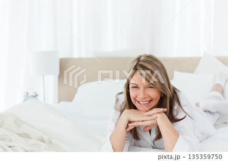 Senior woman lying on bed in bedroom resting chin on hands smiling through sheer curtains Senior woman lying on bed in bedroom resting chin on hands smiling through sheer curtains 135359750
