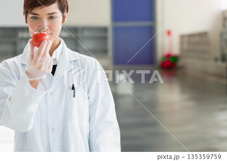 Female researcher wearing white lab coat, gloves inspecting red tomato in lab corridor, copy space Female researcher wearing white lab coat, gloves inspecting red tomato in lab corridor, copy space 135359759