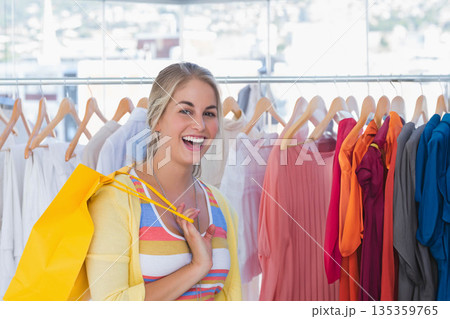 Female shopper standing before metal clothing rack at store holding yellow shopping bag, copy space Female shopper standing before metal clothing rack at store holding yellow shopping bag, copy space 135359765
