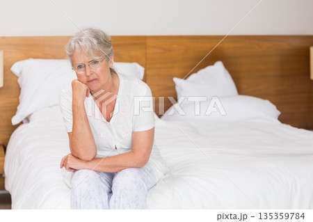 Senior woman resting chin on hand and gazing in bedroom with white bedding, wooden headboard, lamps 135359784