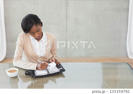 African American woman sitting at glass table writing planner with pen near coffee cup, copy space 135359792