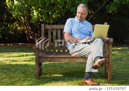 Senior man in polo shirt and khaki trousers sitting on wooden bench in garden using laptop Senior man in polo shirt and khaki trousers sitting on wooden bench in garden using laptop 135359797