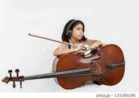 Asian girl in black dress playing cello and shouting with headphone. Concept for music education, child prodigy, emotional performance, and creative talent Asian girl in black dress playing cello and shouting with headphone. Concept for music education, child prodigy, emotional performance, and creative talent 135360290