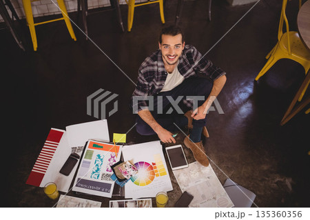 Man sitting cross legged on floor in design studio using smartphone amid design prints and juice 135360356
