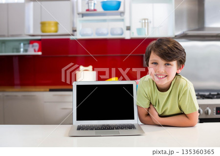 Child boy leaning on countertop and smiling in modern kitchen with silver laptop, copy space Child boy leaning on countertop and smiling in modern kitchen with silver laptop, copy space 135360365