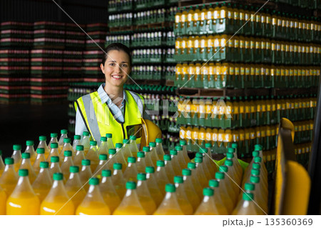 Female warehouse worker holding helmet wearing safety vest in warehouse among beverage bottles 135360369