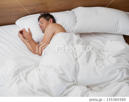 Man sleeping on his side under white duvet in bedroom with wooden headboard, copy space 135360380