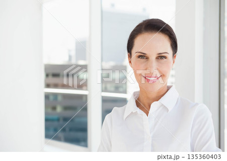 Middle-aged woman wearing white shirt standing before large glass windows in office, copy space 135360403